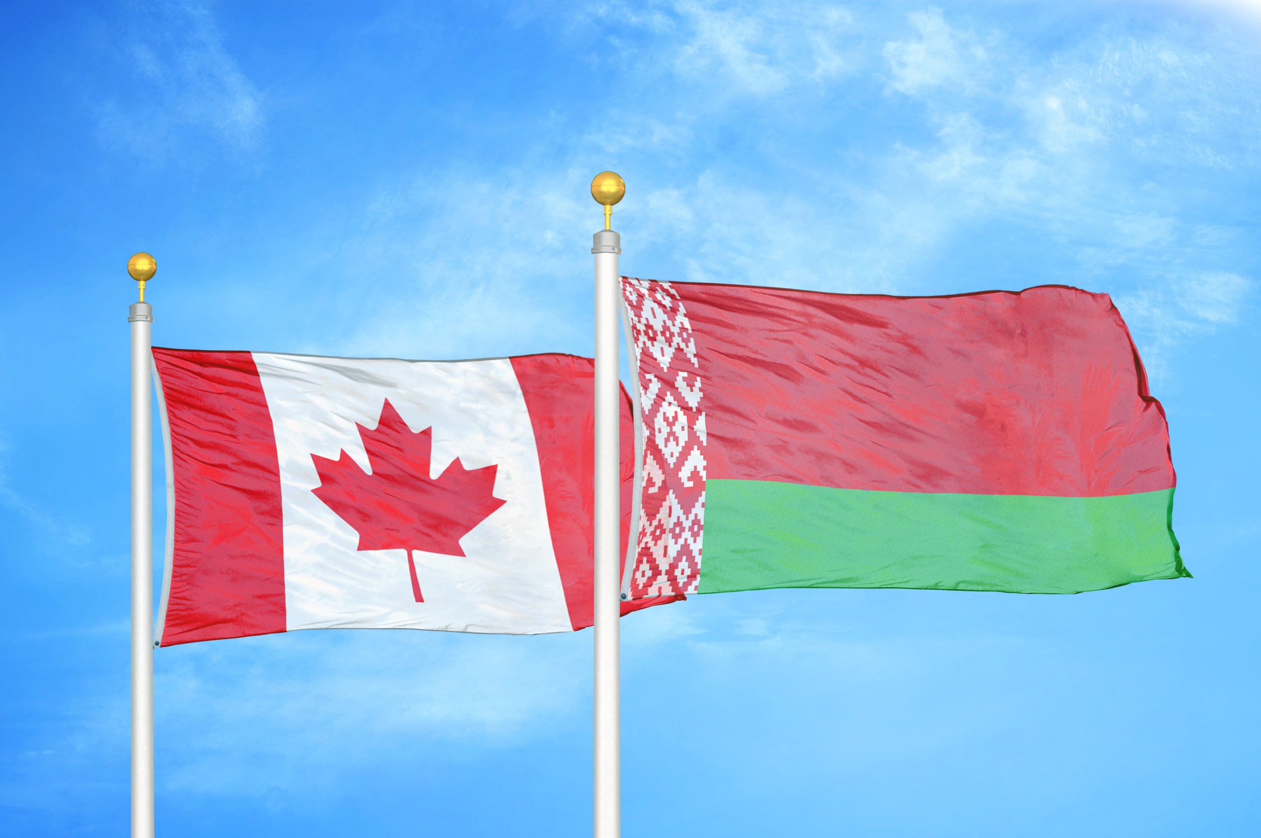 Canada And Belarus Two Flags On Flagpoles And Blue Cloudy Sky Background